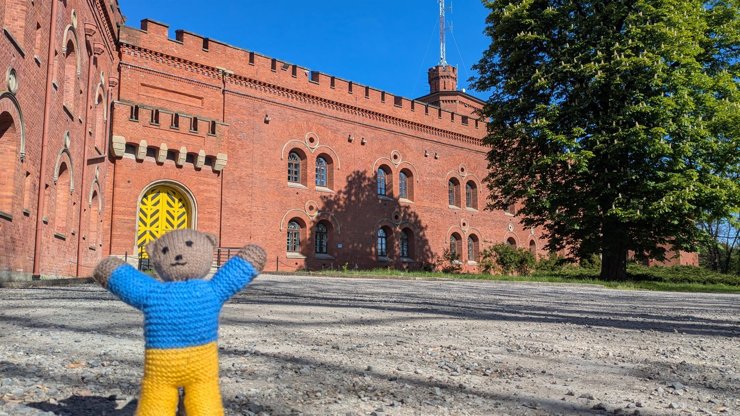 Petya, a hand-knitted teddy bear in a Ukrainian blue-and-yellow jumper, arms raised in front of a red-brick fortress